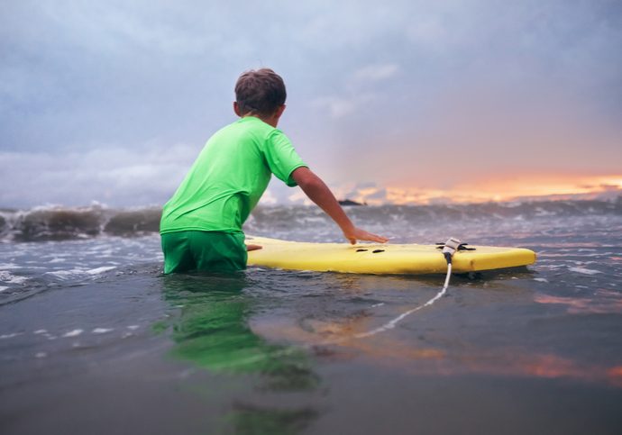 Boy learning to surf in ocean waves at sunset time